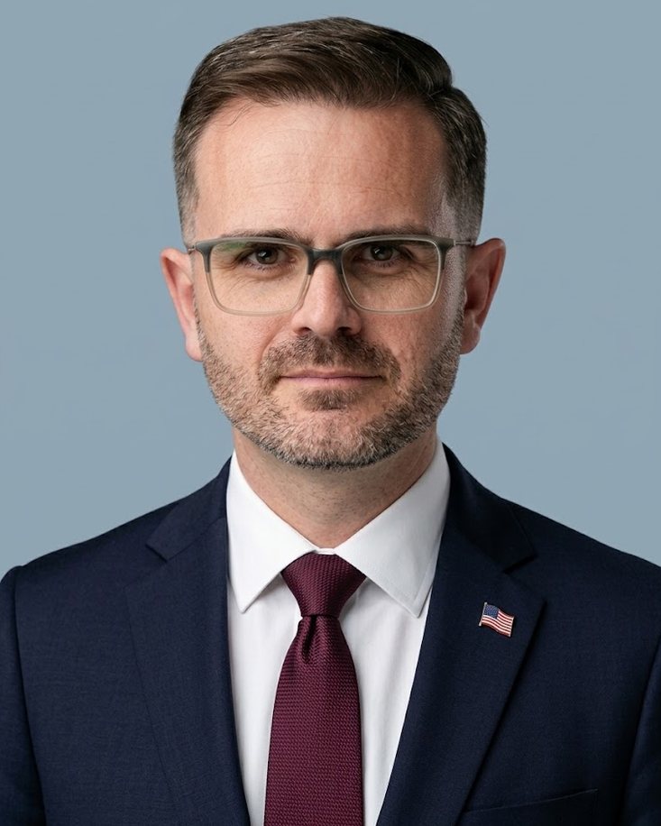 Mark Malinowski in a navy suit, white shirt, and burgundy tie, against a light blue background